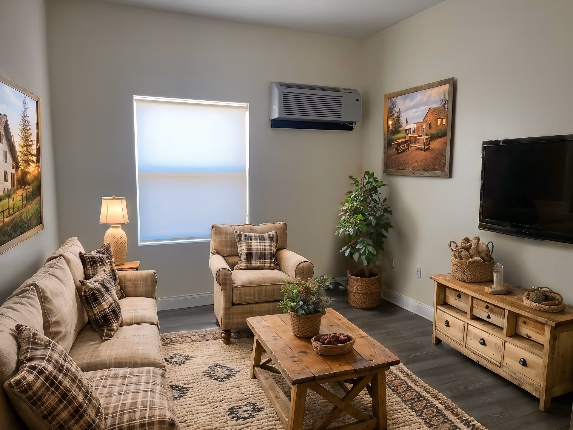 A cozy living room with a beige sofa and armchair featuring plaid cushions. A wooden coffee table with a plant and a bowl of fruit sits on a patterned rug. The room has a window with a shade, a wall-mounted air conditioner, a flat-screen TV on the wall, a wooden media console with baskets and decor, and two framed pictures on the walls. A potted plant is placed in the corner.