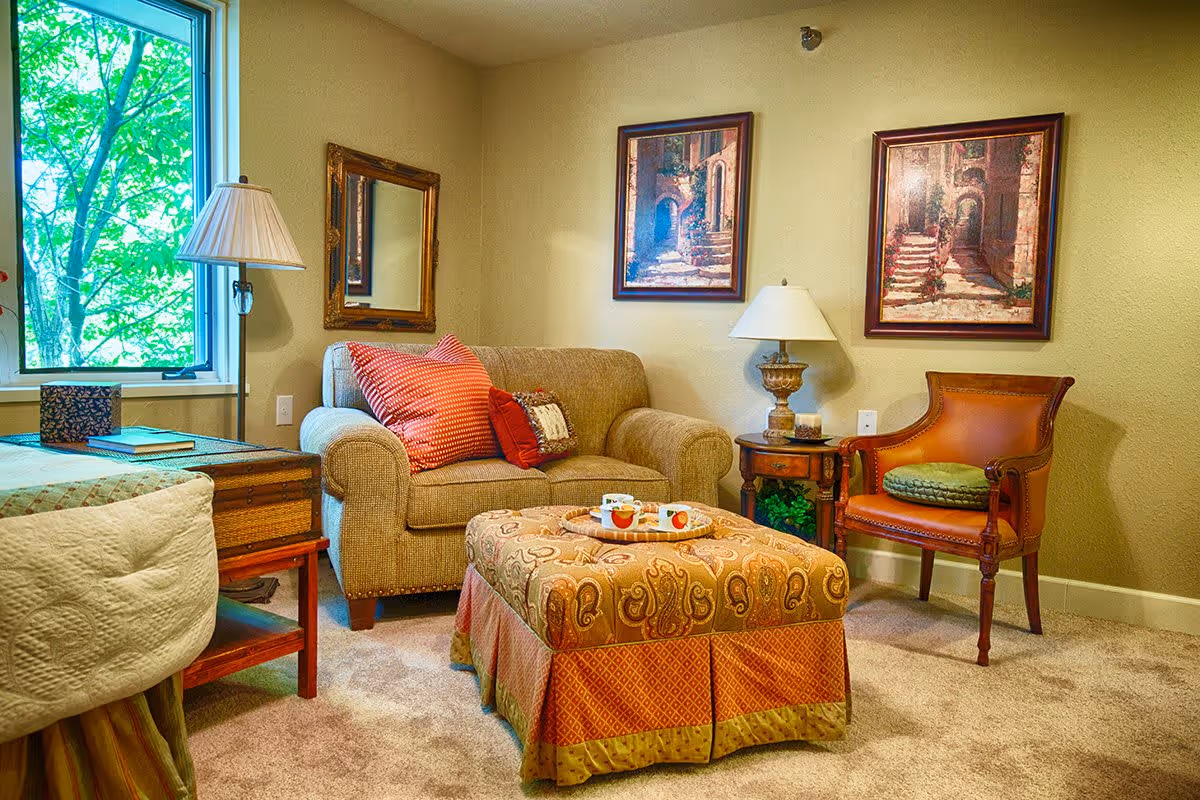 A cozy living room area with a beige loveseat adorned with red and patterned throw pillows, a large patterned ottoman with a tray holding two cups, a wooden side table with a lamp and candle, a leather armchair with a green cushion, two framed paintings on the wall, a floor lamp next to a window showing green tree foliage outside, and a bed partially visible on the left side.