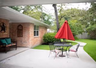 Outdoor patio area with a round metal table and four chairs, each with a cushion. A red umbrella is closed and positioned in the center of the table. The patio is adjacent to a brick building with a window and some outdoor seating with cushions. There are trees and green grass surrounding the patio area.