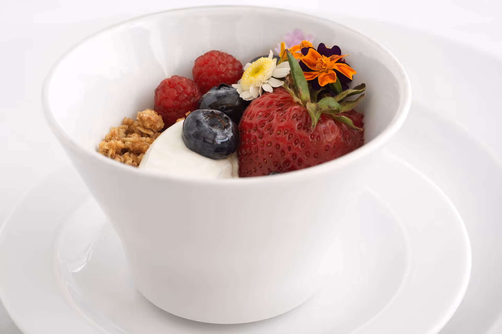 A white bowl on a white plate containing granola, raspberries, blueberries, a strawberry, a dollop of yogurt, and small edible flowers.