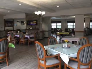 A spacious dining area with multiple tables and chairs arranged neatly. Each table has a tablecloth and a small centerpiece with flowers. The room features large windows allowing natural light, a fireplace with a mounted TV above it, and a chandelier hanging from the ceiling. The flooring is wood-style, and the overall atmosphere is warm and inviting.