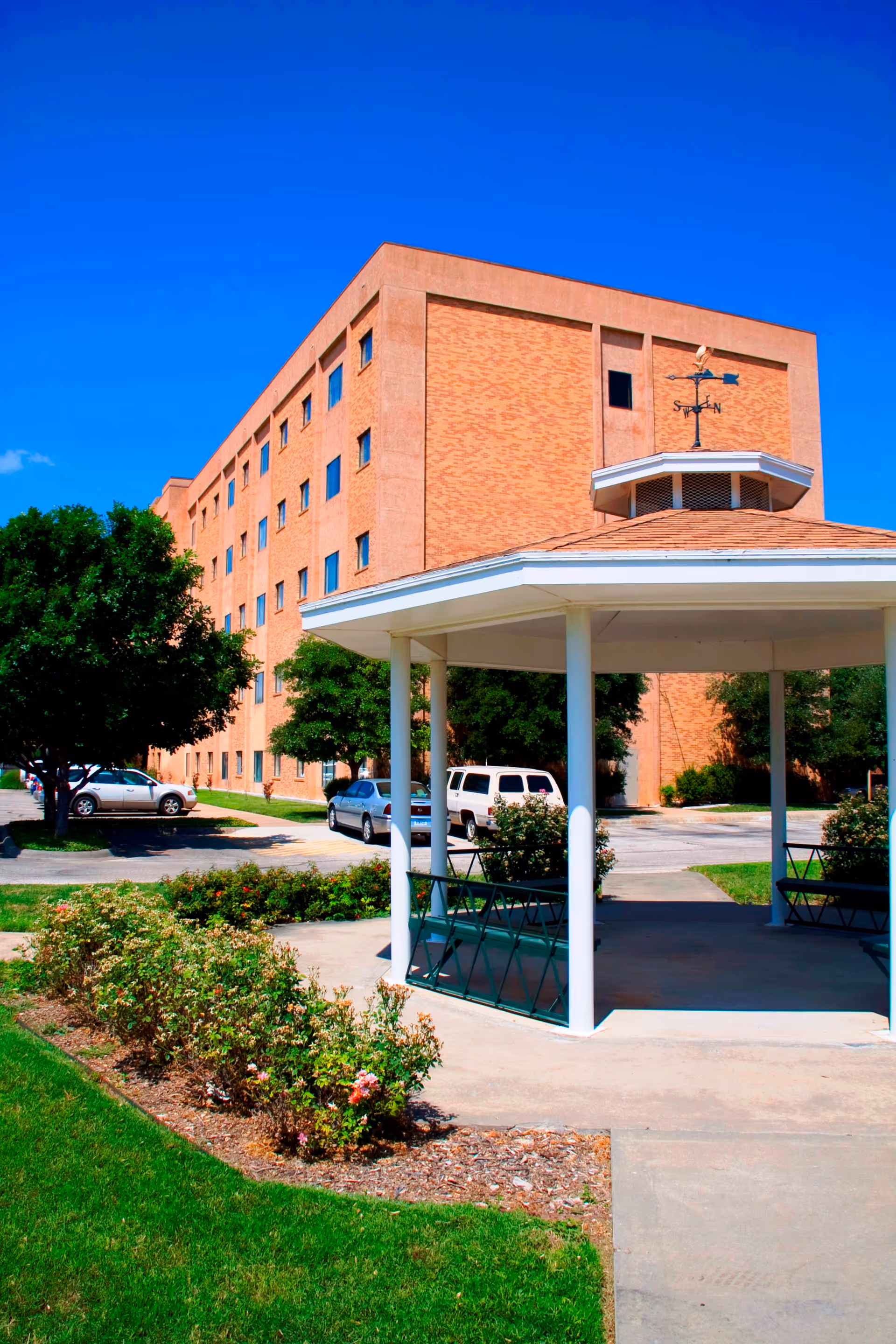 Brick multi-story senior living building with a white-roofed gazebo, parked cars, and landscaped grounds under a clear blue sky.