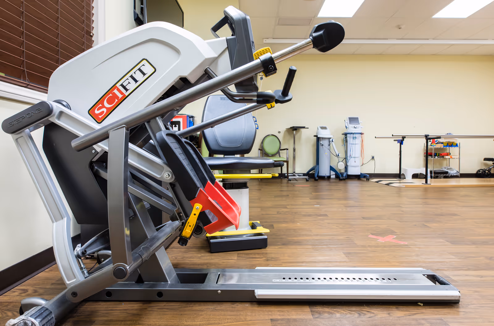 A SciFit exercise machine in a room with wooden flooring and beige walls, featuring other medical and rehabilitation equipment in the background.
