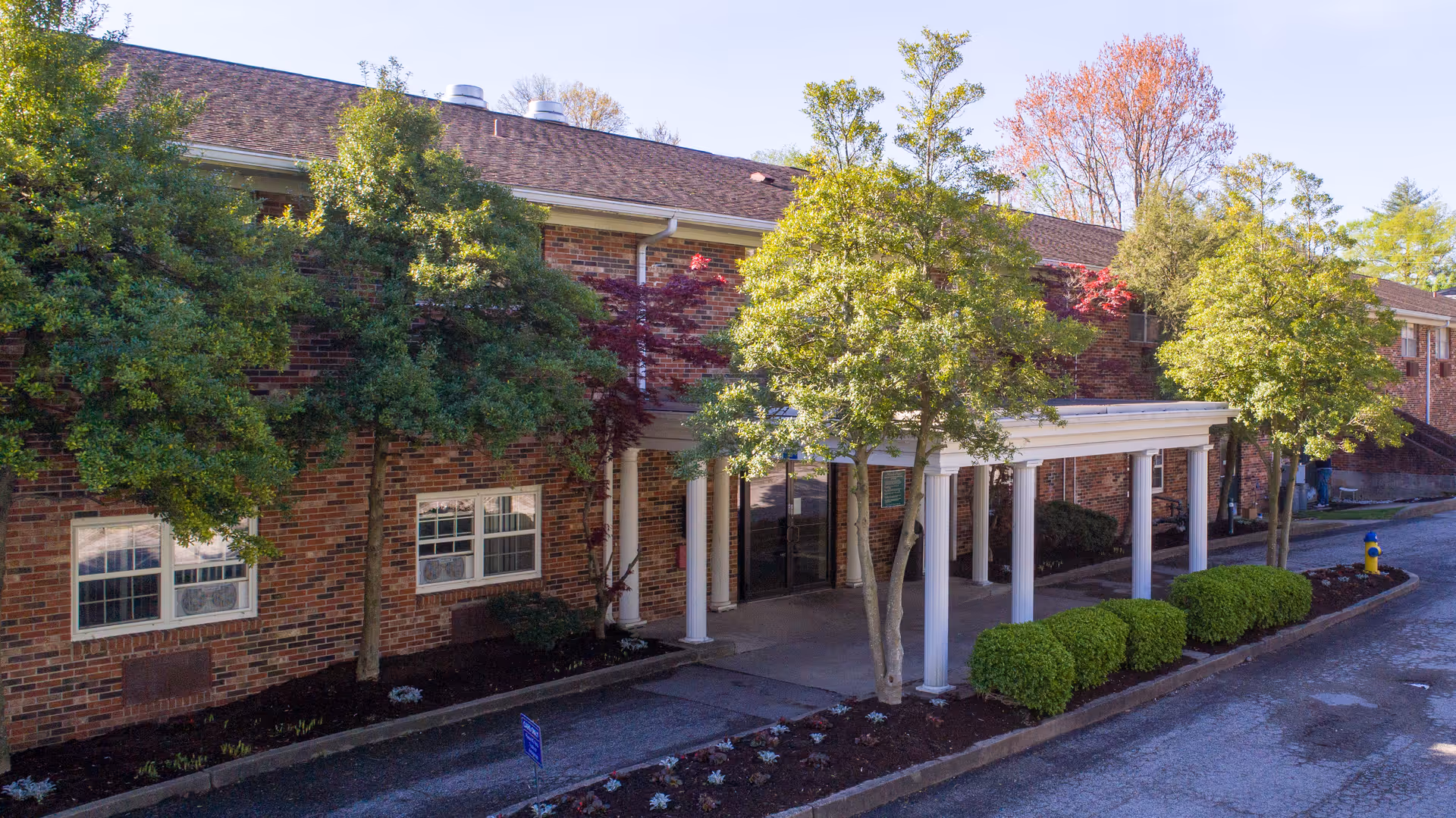 Exterior view of a brick building with a covered entrance supported by white columns. There are several trees and bushes along the walkway leading to the entrance. The sky is clear and the scene is well-lit by daylight.