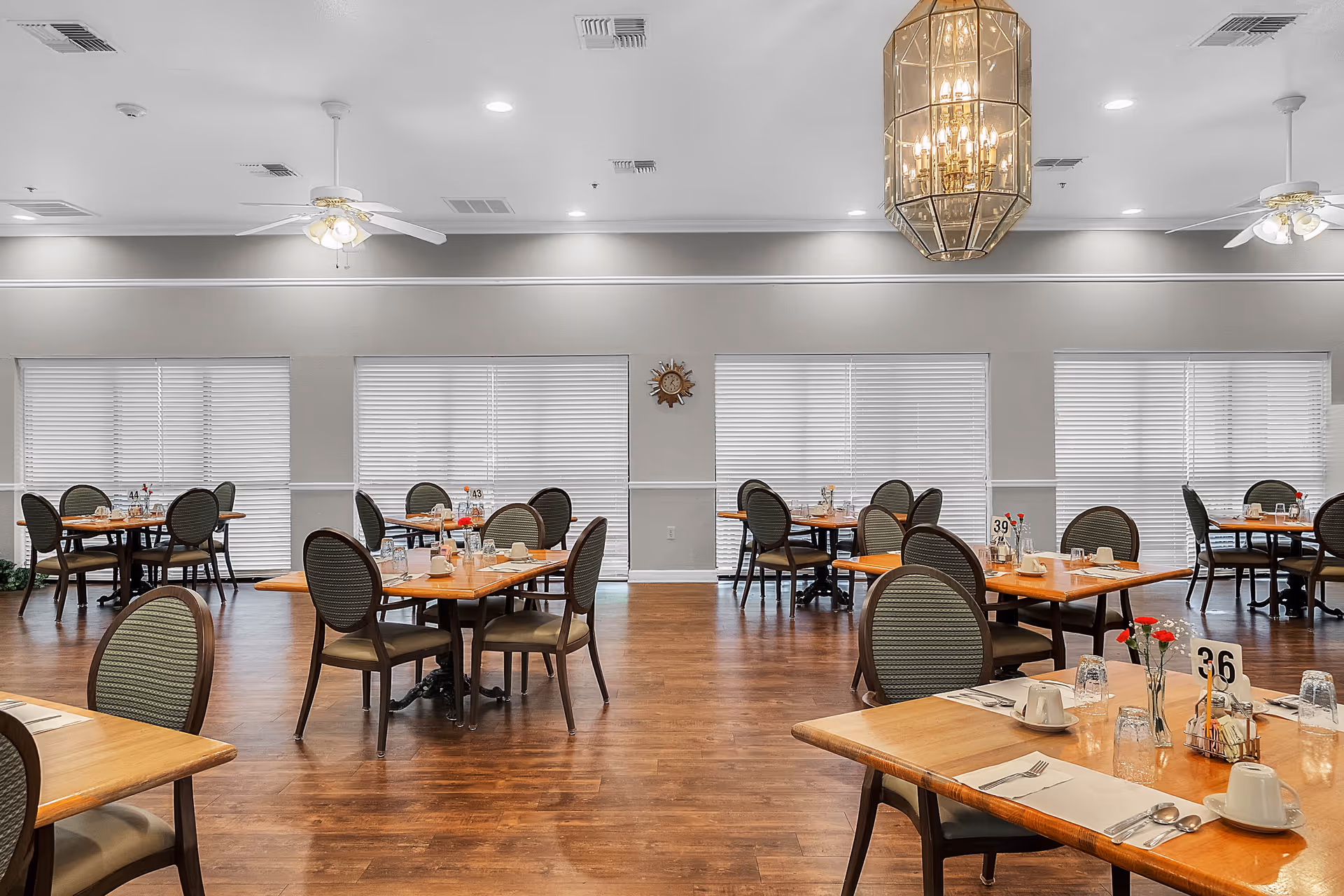 A spacious dining room with multiple wooden tables and cushioned chairs arranged neatly. Each table is set with placemats, cups, glasses, and silverware. The room has large windows with white blinds, ceiling fans, recessed lighting, and a large decorative chandelier hanging from the ceiling. The floor is wooden, and the walls are painted light gray with white trim.