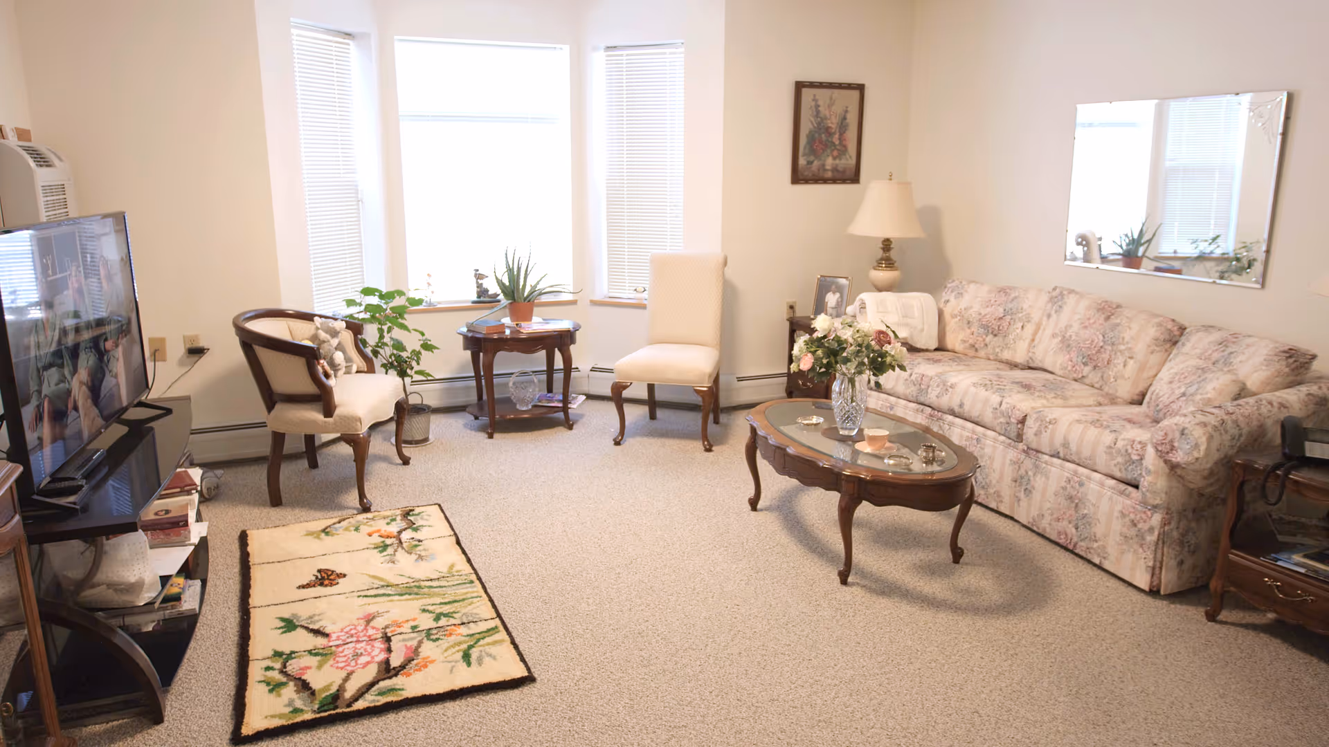 A cozy living room with a floral-patterned sofa, a glass-top wooden coffee table with a vase of flowers, two upholstered chairs, a small round wooden side table with plants, a flat-screen TV on a stand, and a decorative rug on a carpeted floor. The room has three large windows with blinds and a wall mirror above the sofa.