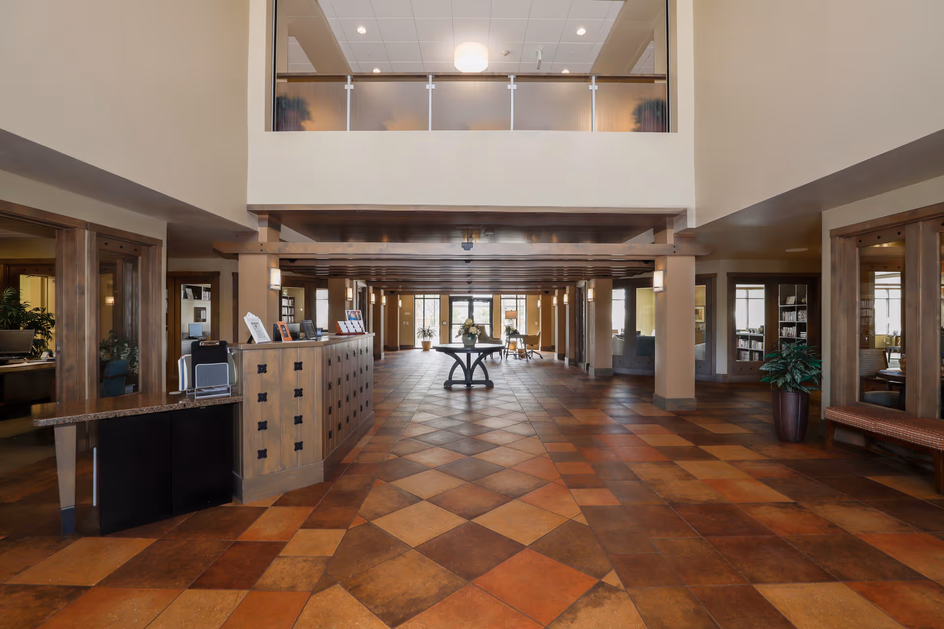 Spacious lobby area with a high ceiling and tiled floor in warm tones. There is a wooden reception desk on the left side, a round table with a flower arrangement in the center, and seating areas with plants and bookshelves visible in the background. The upper level has a glass railing overlooking the lobby.