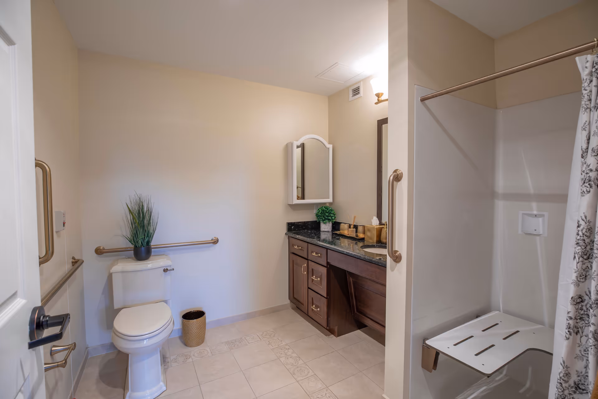 A clean and accessible bathroom with beige walls and tiled floor. The room features a white toilet with a potted plant on top, grab bars on the walls, a wooden vanity with a black countertop, a mirror, and a light fixture above. There is also a shower area with a fold-down seat and a floral-patterned shower curtain.