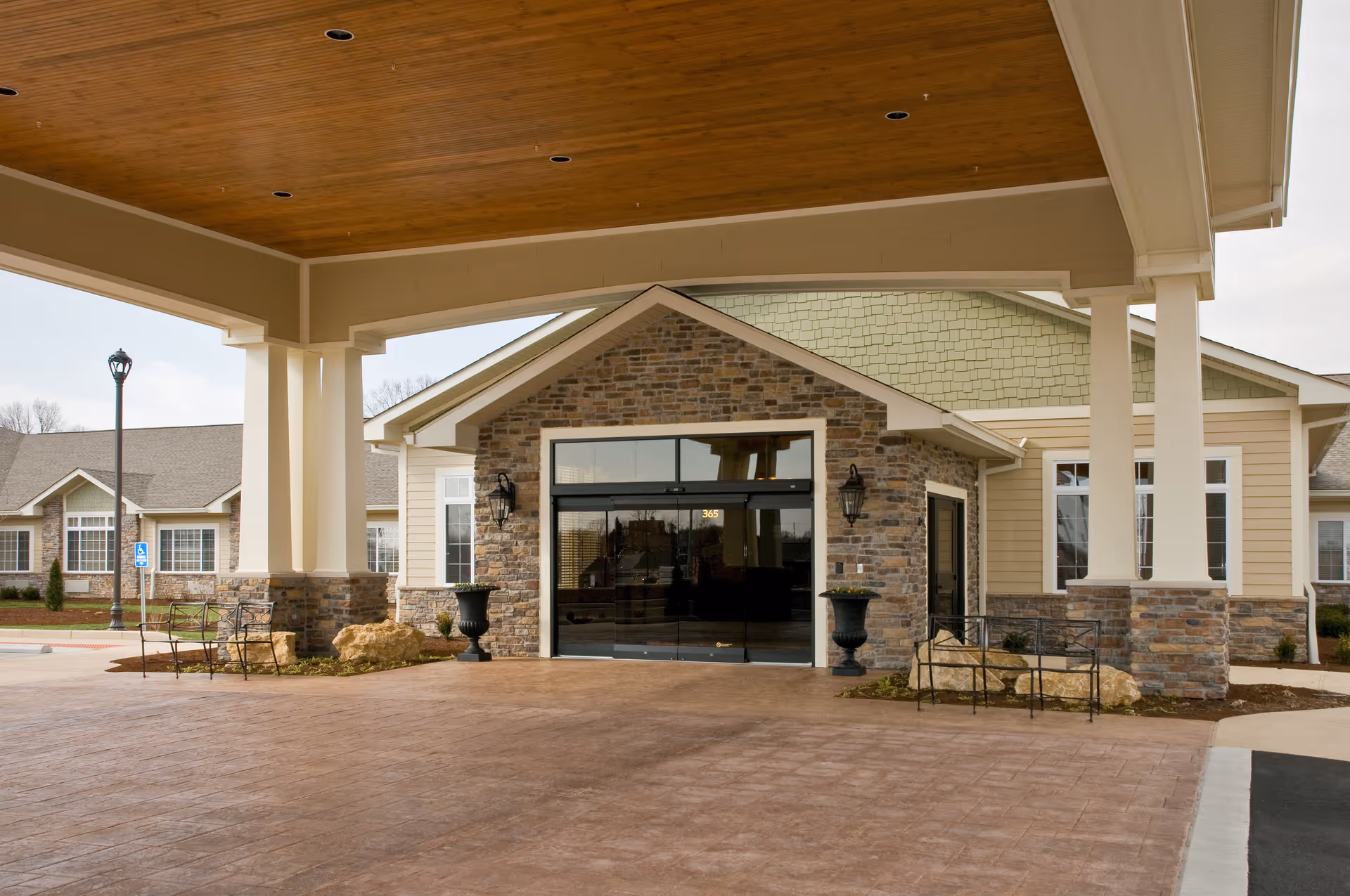 Covered entrance area of a senior living facility with stone and beige siding exterior walls, large glass double doors, two black urn planters, and metal benches on either side. The building has a green shingled roof and a wooden ceiling under the covered area.