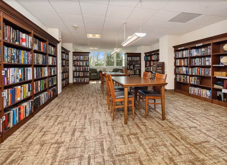 A bright interior reading room with bookshelves along the walls, a central table with chairs, and seating by a window.