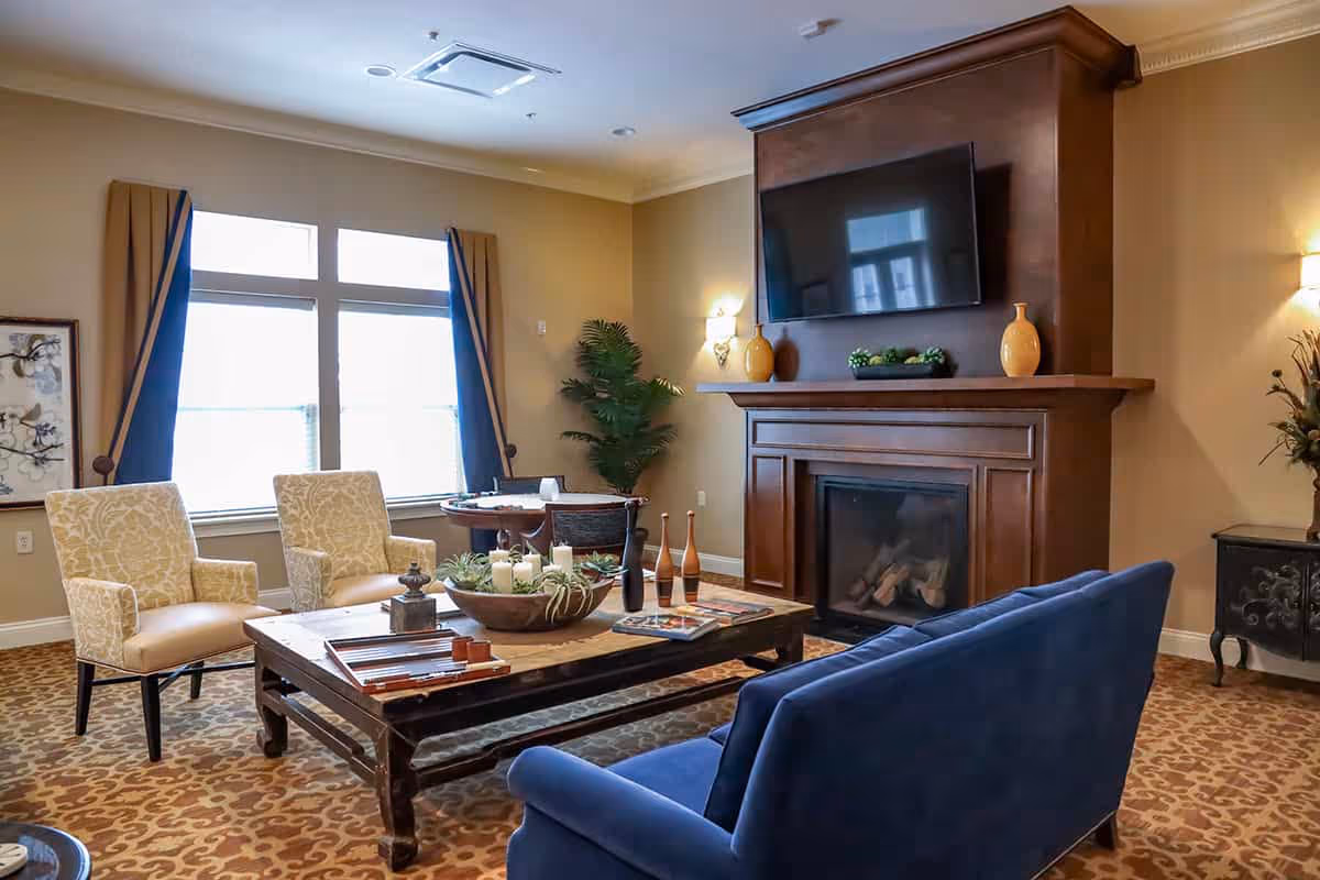 Well-appointed living room with upholstered chairs and a blue sofa around a wooden coffee table, a fireplace with a wall-mounted TV, and large windows.