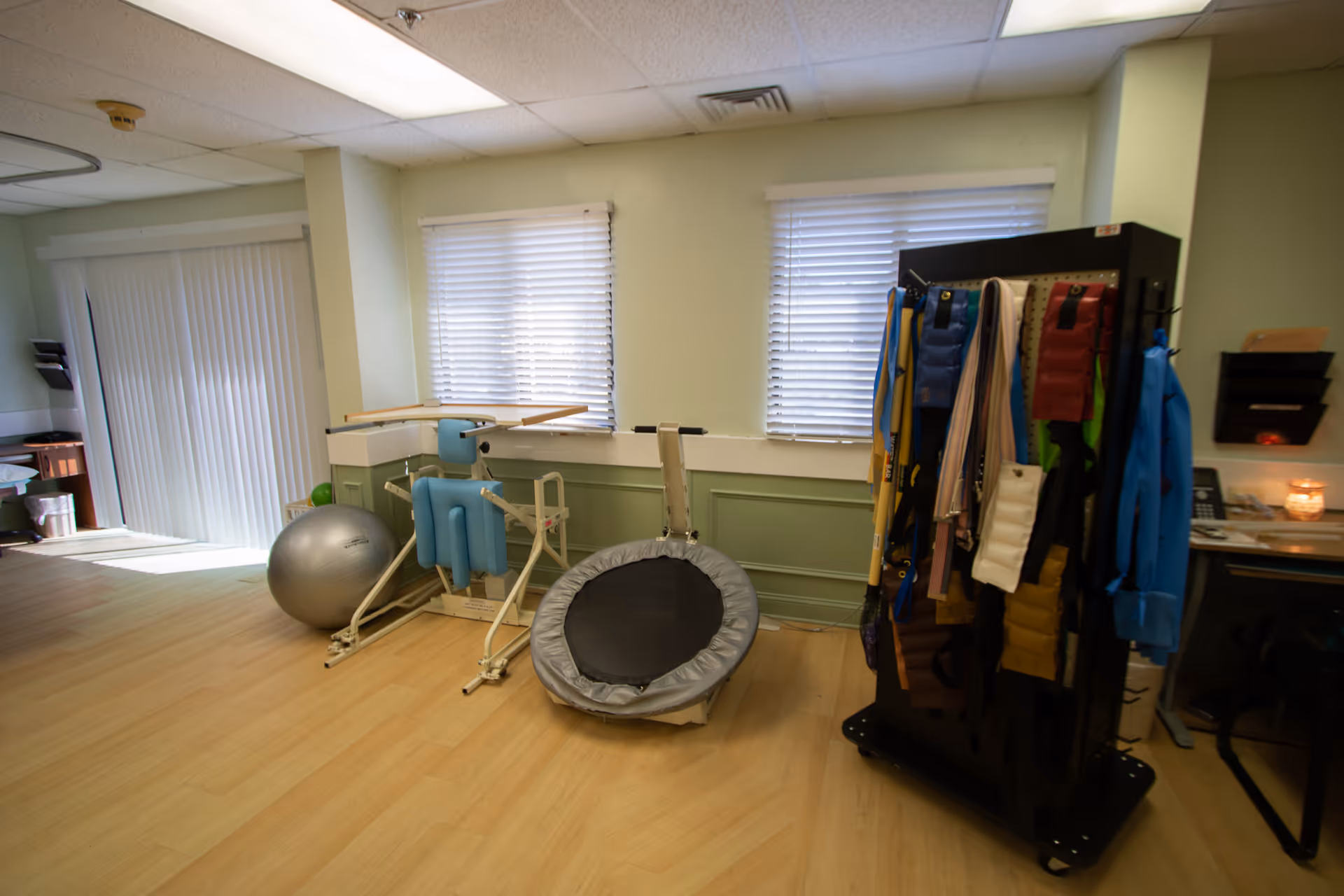 Therapy/rehab room with exercise equipment including an exercise ball, mini-trampoline, gait belts and parallel bars beneath two windows.