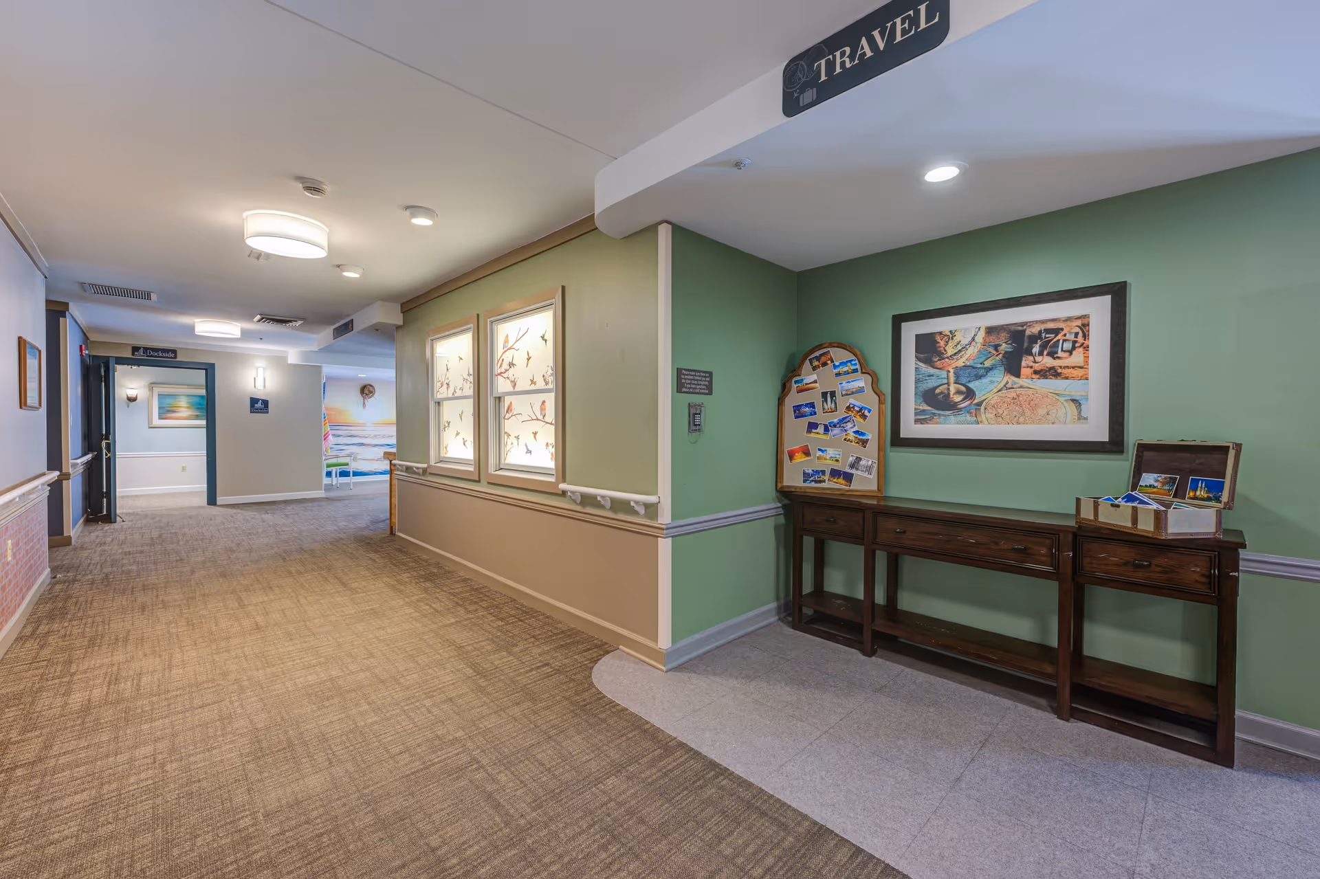 A well-lit hallway in a senior living facility with carpeted flooring and beige and green walls. On the right side, there is a wooden console table with postcards and a framed travel-themed picture above it. A sign labeled 'TRAVEL' hangs from the ceiling above the table. The hallway extends into a seating area with colorful wall art visible in the background.