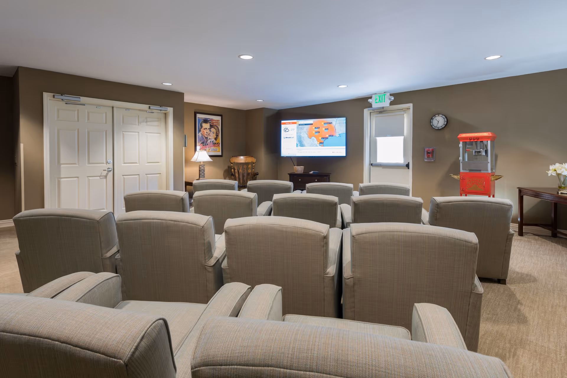 Small screening room with rows of upholstered chairs facing a wall-mounted TV, a popcorn machine, and an exit door.