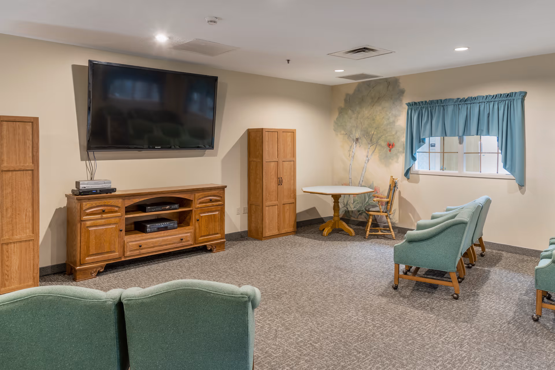 A cozy common room with a wall-mounted TV, wooden media cabinet, green upholstered chairs, and a small table by a window.
