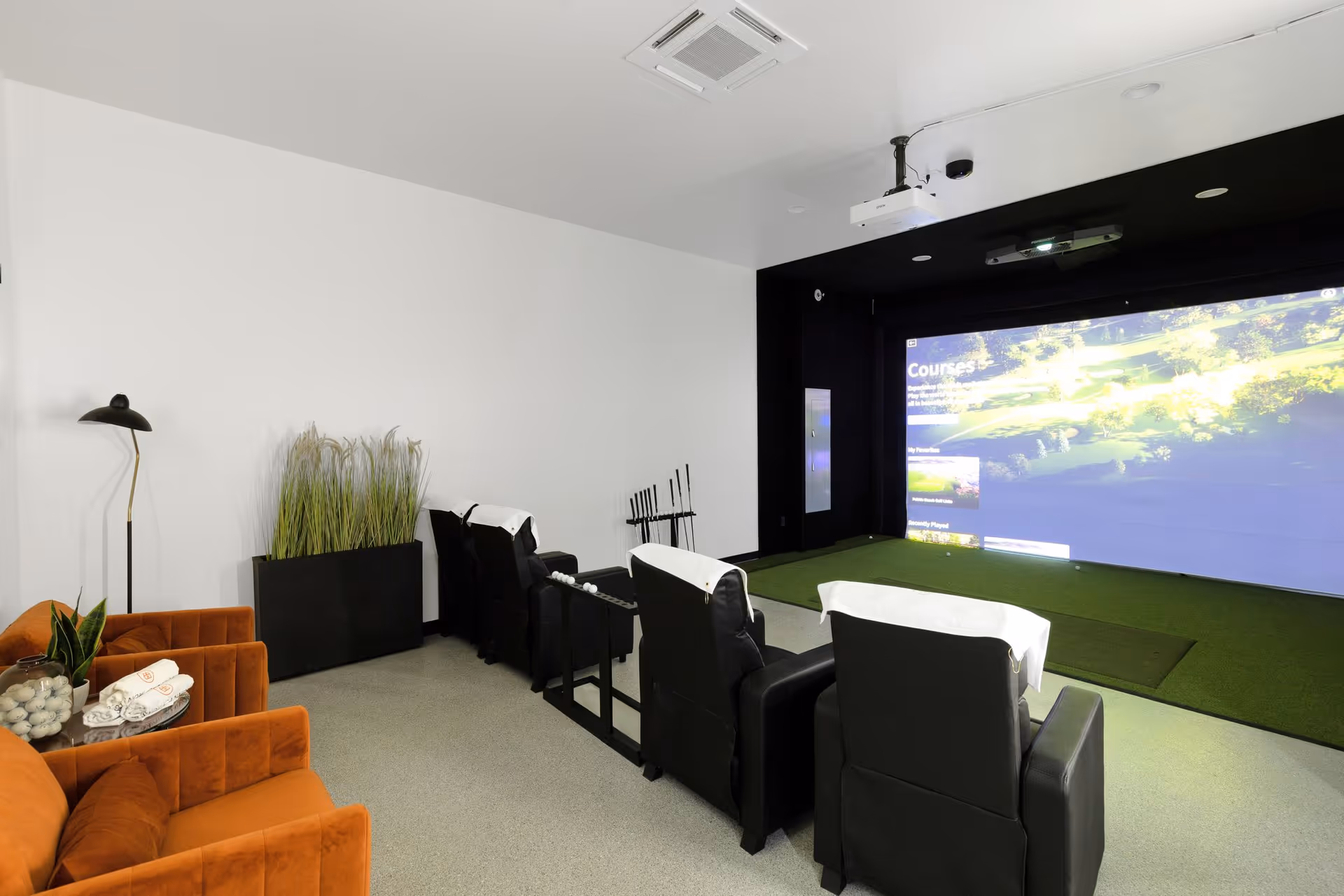 Indoor golf simulator room with recliner seating facing a large projection screen and putting turf.