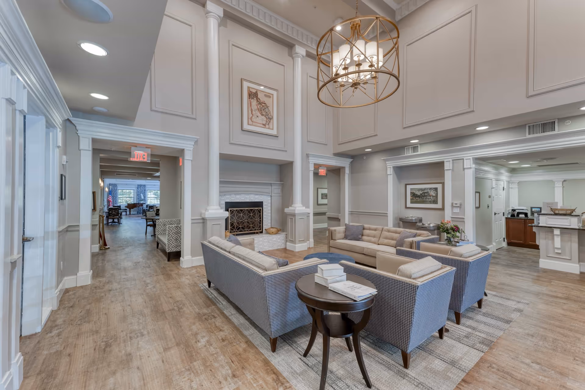 A spacious and elegant living room area in a senior living facility with high ceilings, a large chandelier, and a fireplace. The room features comfortable seating including sofas and armchairs arranged around a coffee table with books. The walls are decorated with framed artwork, and the flooring is wood. There are doorways leading to other parts of the facility.