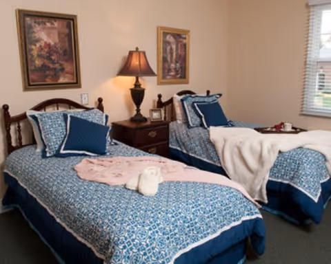 Two twin beds with blue patterned bedding separated by a nightstand with a lamp, framed art on the wall and a window to the right.