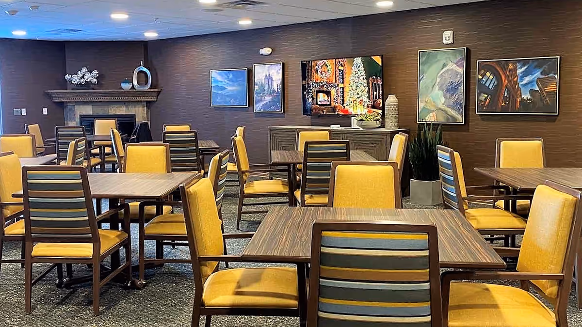 A spacious dining room with multiple wooden tables and yellow cushioned chairs. The room features a fireplace on the left wall, several framed pictures and a flat-screen TV mounted on the back wall, and decorative plants. The ceiling has recessed lighting, and the overall ambiance is warm and inviting.
