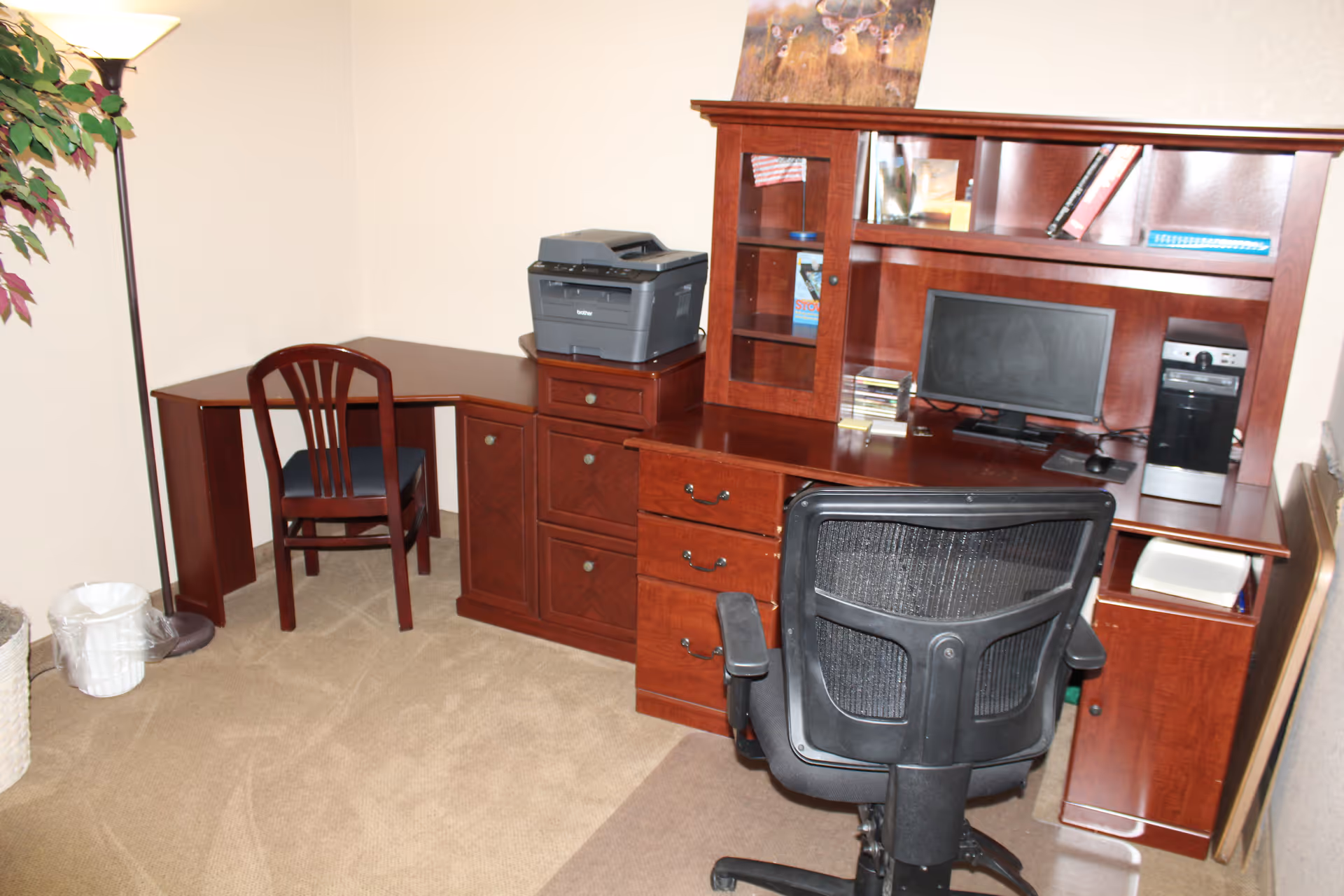 An office corner in a room with beige walls and carpeted floor. The office setup includes a dark wooden L-shaped desk with multiple drawers and cabinets. On the desk, there is a computer monitor, a desktop computer tower, a printer, and some books and decorative items on the shelves. There are two chairs: a black mesh office chair and a wooden chair with a cushioned seat. A tall floor lamp and a potted plant are visible in the corner.