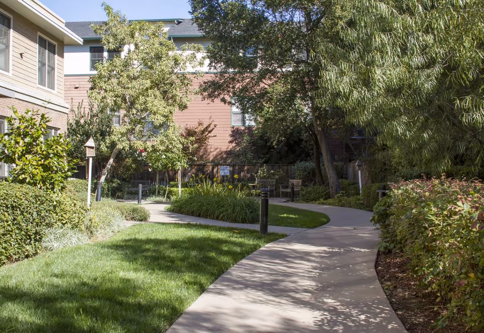 A curved concrete pathway winds through a well-maintained garden area with green grass, bushes, and trees. There are benches along the path and a building with windows visible in the background under a clear blue sky.