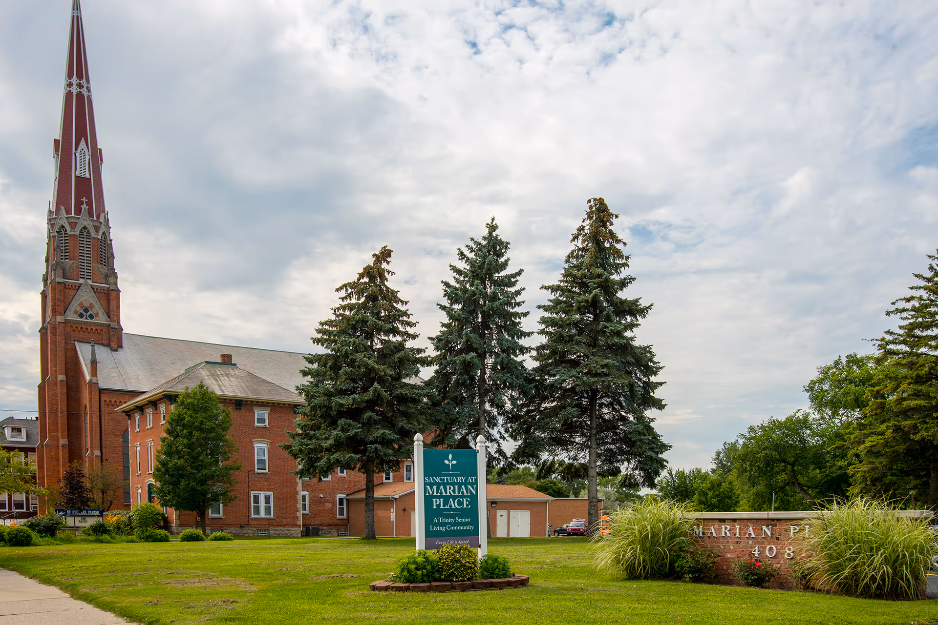 Exterior view of Marian Place senior living community featuring a large brick building with a tall church steeple, several evergreen trees, a green sign that reads 'Sanctuary at Marian Place A Trinity Senior Living Community,' and a brick sign with the address number 408 on a well-maintained grassy lawn under a partly cloudy sky.