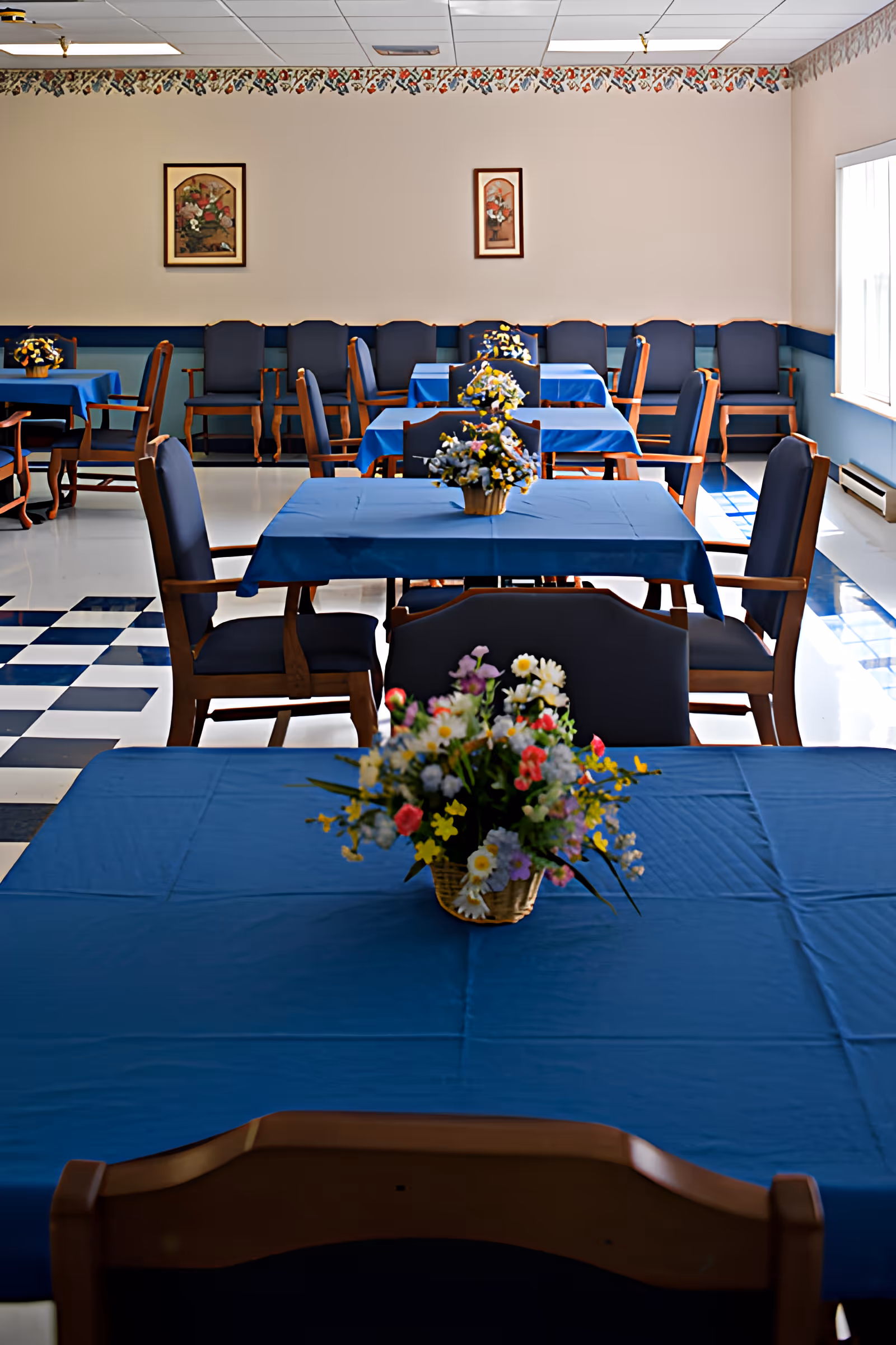 A dining room with multiple tables covered in blue tablecloths, each adorned with a floral centerpiece. The room has wooden chairs with dark cushions, a checkered black and white floor, and framed floral artwork on the walls. Natural light enters through a window on the right side.