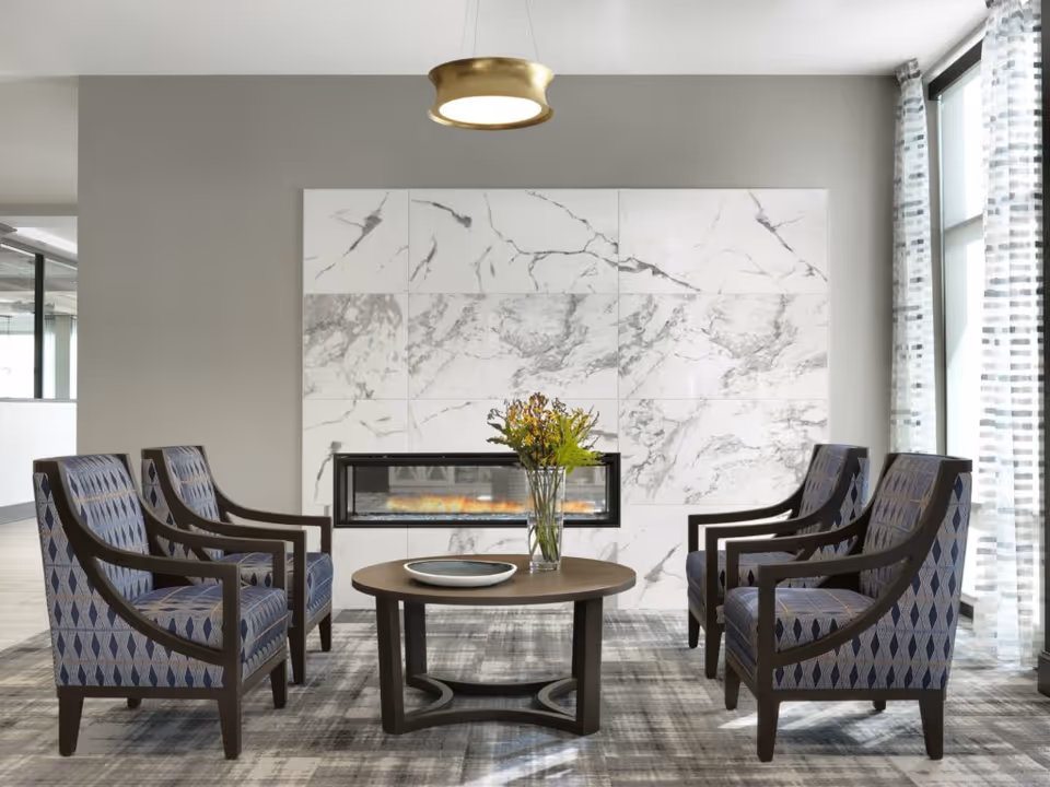 A modern sitting area with four patterned armchairs arranged around a round wooden coffee table with a vase of flowers and a decorative plate. Behind the seating is a wall with large white marble tiles and a built-in horizontal electric fireplace. To the right, tall windows with sheer patterned curtains allow natural light into the room.