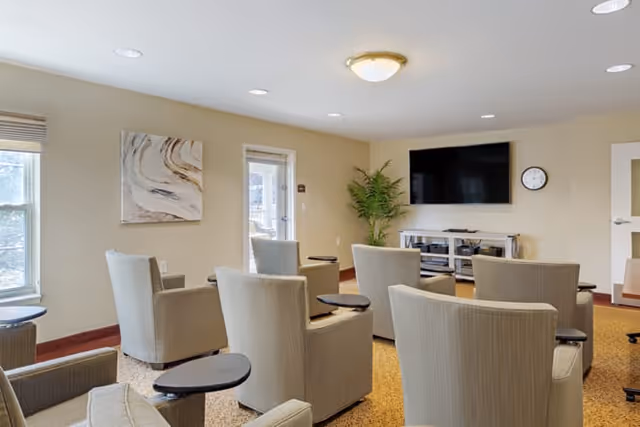 A small common area with several beige armchairs arranged in rows facing a wall-mounted flat screen TV. The room has beige walls, a large window, a door with glass panels, a potted plant, a wall clock, and a piece of abstract artwork on the wall.