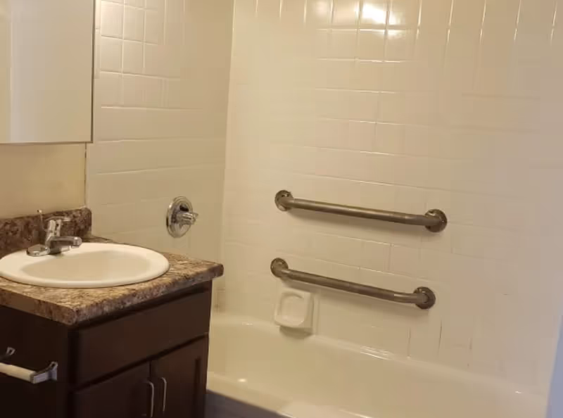Bathroom vanity with sink next to a tiled bathtub equipped with two horizontal grab bars.