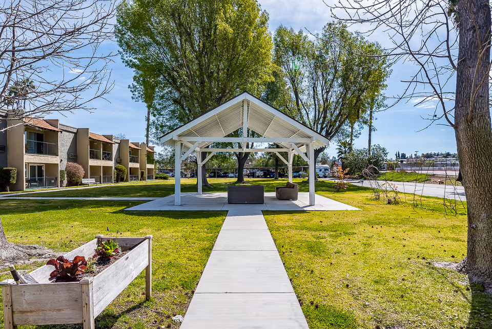 A white covered pavilion with seating in a grassy courtyard surrounded by apartment buildings and trees.