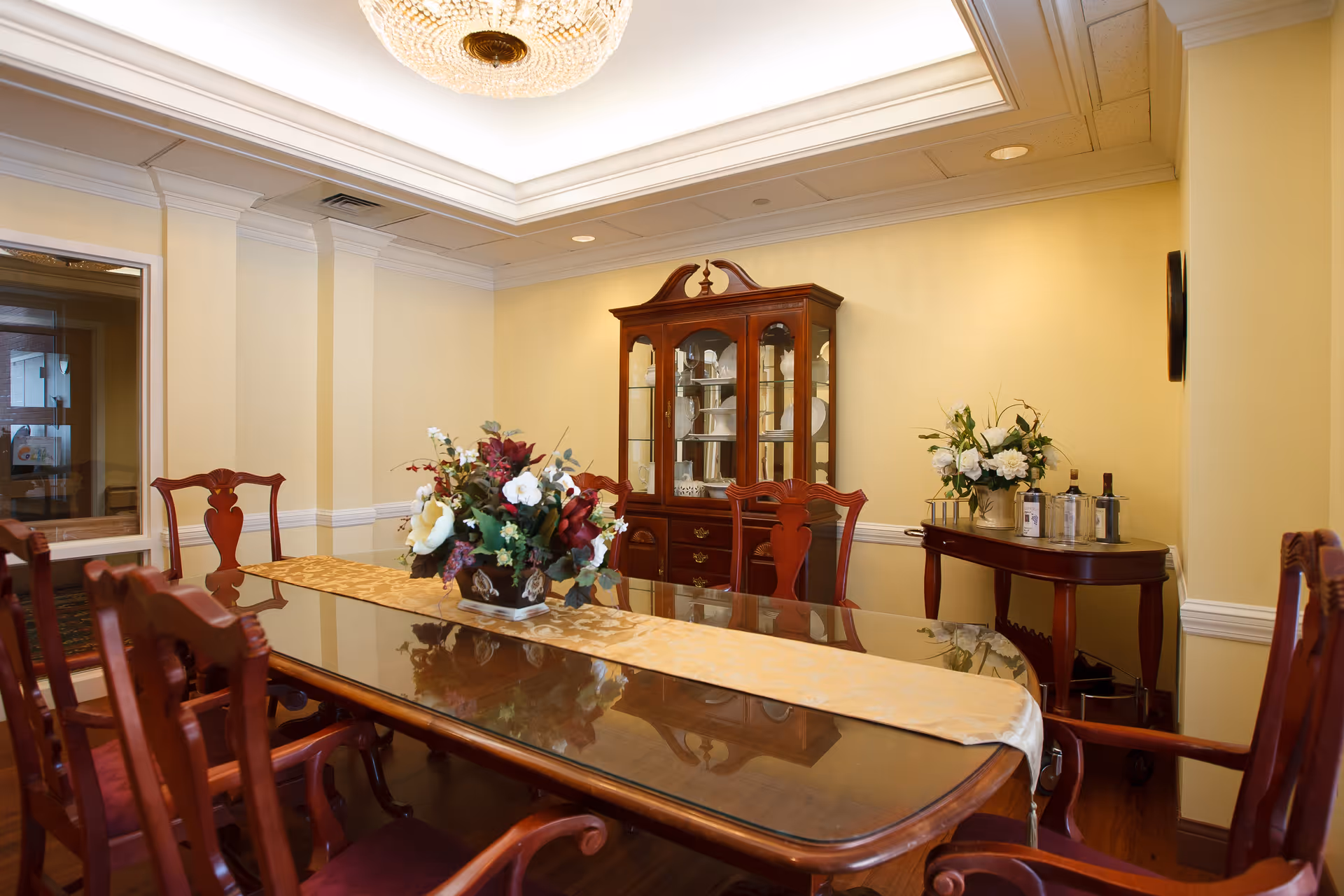 A formal dining room with a polished wooden table and six matching chairs. The table has a beige runner and a floral centerpiece. Against the wall is a wooden china cabinet displaying plates and glassware. To the right, a small wooden side table holds a flower arrangement and several bottles. The room has yellow walls, crown molding, and a decorative ceiling light fixture.