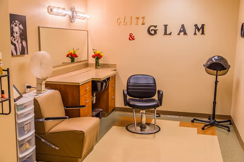 Interior view of a small salon room with two salon chairs, a hair dryer, a countertop with a mirror, and a vase with flowers. The wall has decorative letters spelling 'GLITZ & GLAM' and a black and white photo of a woman.