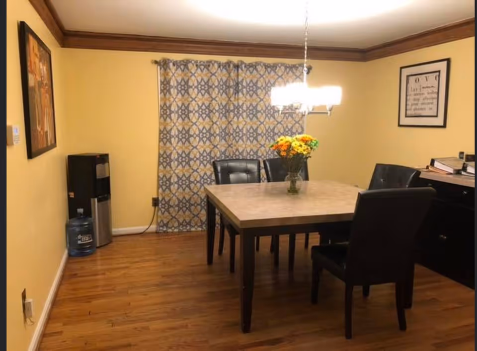 A dining room with a square table and four black cushioned chairs. A vase with colorful flowers is placed on the table. The room has yellow walls, wooden flooring, and a patterned curtain covering a window. There is a water dispenser with a large water bottle on the left side and framed artwork on the walls. A chandelier light fixture hangs above the table.