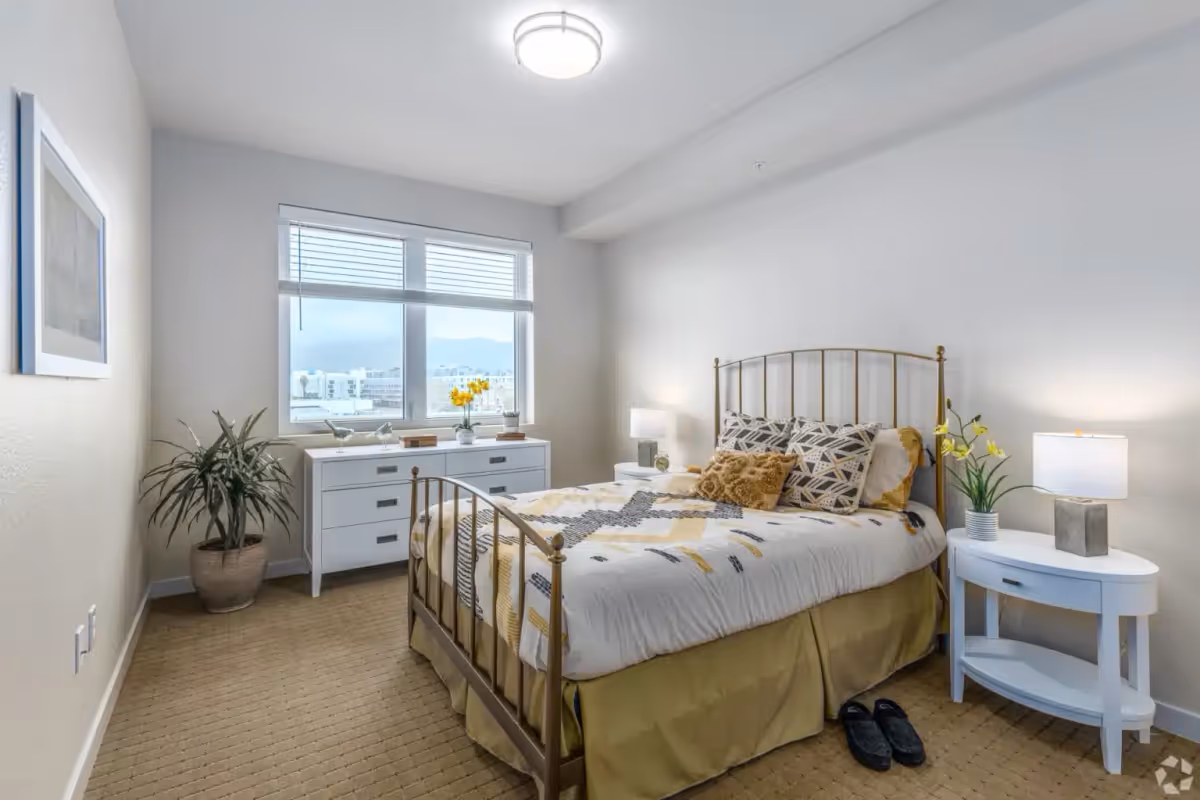 A bright and neatly arranged bedroom with a metal bed frame, patterned bedding, two pillows, and a decorative cushion. There is a white nightstand with a lamp and a potted plant on the right side of the bed. Across from the bed is a white dresser with decorative items and a large window with blinds letting in natural light. A potted plant is placed in the corner near the dresser, and a pair of slippers is on the carpeted floor beside the bed.