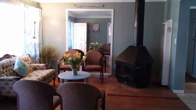 Cozy living room with upholstered chairs and sofa arranged around a small round coffee table with flowers and a black wood-burning stove against a green wall.