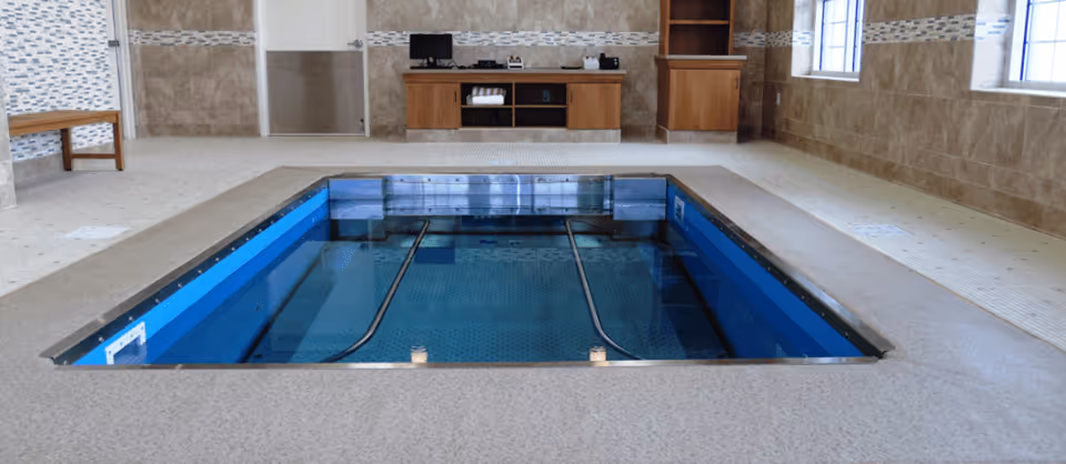 Indoor therapy pool with handrails on both sides, surrounded by beige tiled walls and floor. There is a wooden bench on the left side and a wooden cabinet with electronic equipment against the far wall. Two windows on the right wall allow natural light into the room.