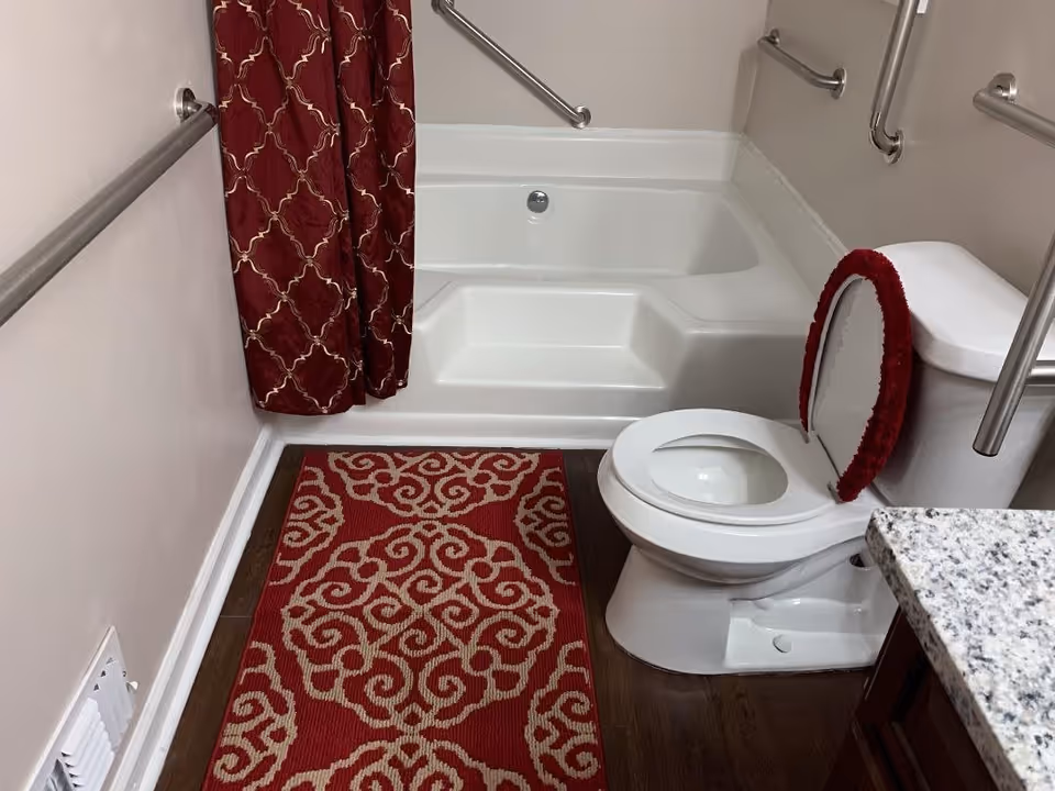 A bathroom featuring a white bathtub with built-in steps and multiple stainless steel grab bars on the walls. There is a white toilet with a red cushioned toilet seat cover and a matching red and beige patterned rug on the dark wooden floor. A red shower curtain with a gold pattern hangs beside the bathtub, and a granite countertop is partially visible on the right side.