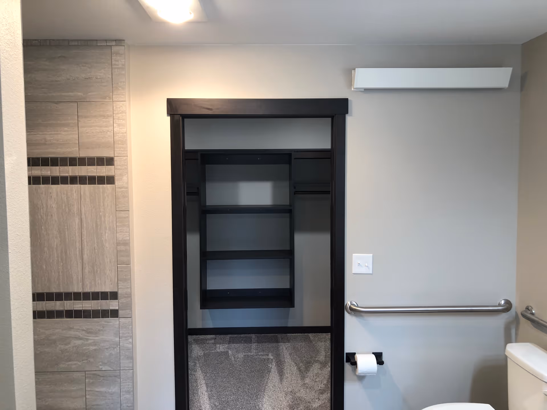 A bathroom with a toilet, grab bars on the wall, a toilet paper holder, and a tiled shower area. There is an open closet with shelves and hanging rods framed in dark wood. The walls are painted light gray.