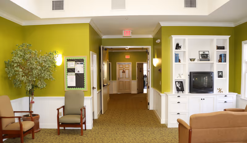 Interior view of a senior living facility common area with green walls, carpeted floor, and white wainscoting. There are chairs arranged along the walls, a potted plant, a bulletin board, and a white built-in shelving unit with a TV and framed photos. A hallway with doors and exit signs is visible in the background.