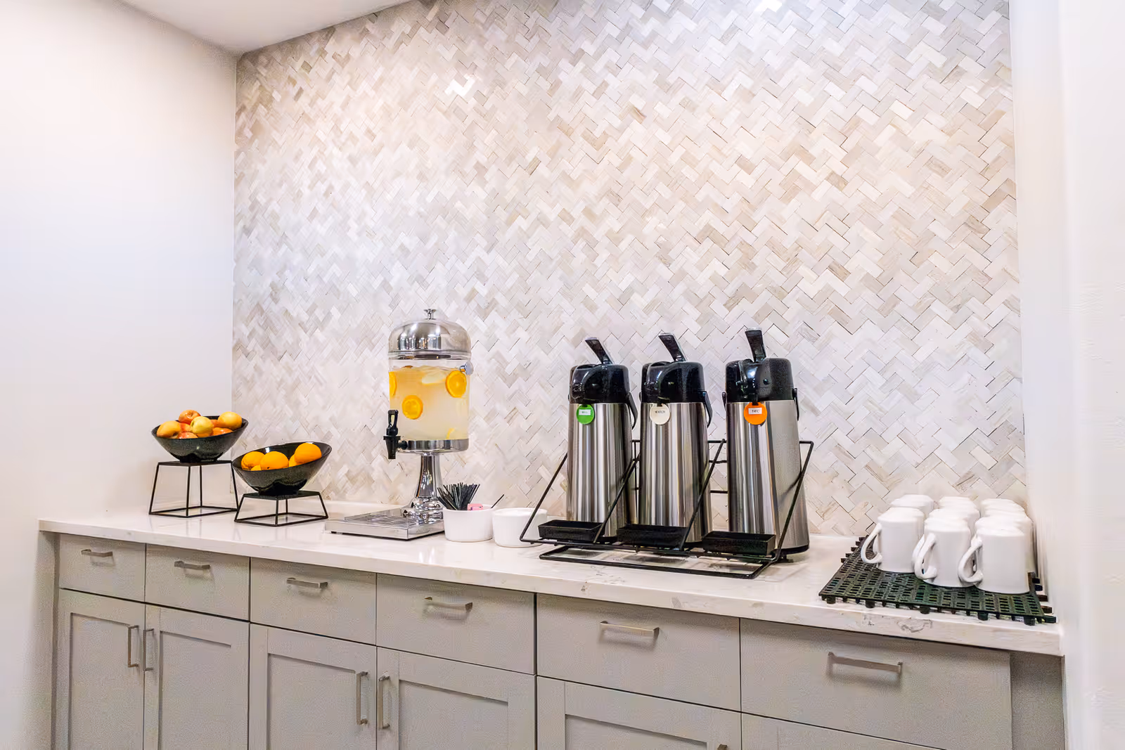 Countertop beverage station with three coffee dispensers, a lemon water dispenser, bowls of fruit, and stacked mugs against a tiled backsplash.