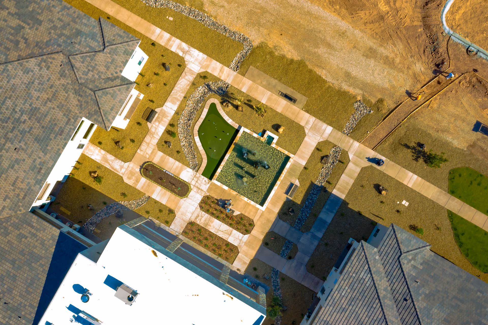 Aerial view of a landscaped courtyard area with walkways, a small pond with fountains, benches, and surrounding buildings with tiled roofs.