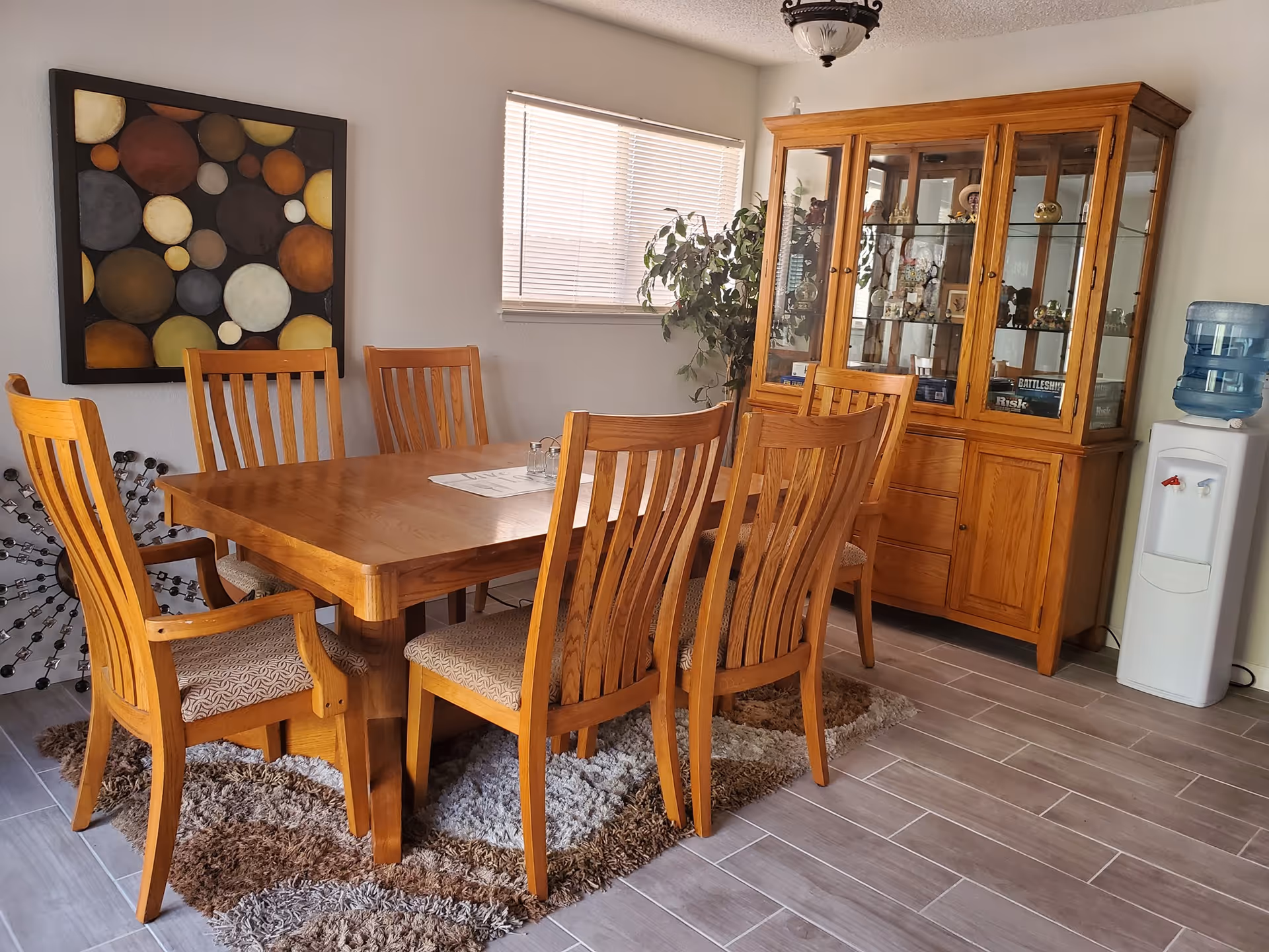 A dining room with a wooden table surrounded by six wooden chairs with cushioned seats. There is a large wooden cabinet with glass doors displaying various decorative items. A water cooler is placed to the right of the cabinet. A window with blinds lets in natural light, and a colorful abstract painting hangs on the wall to the left. The floor is tiled with a patterned rug under the table.