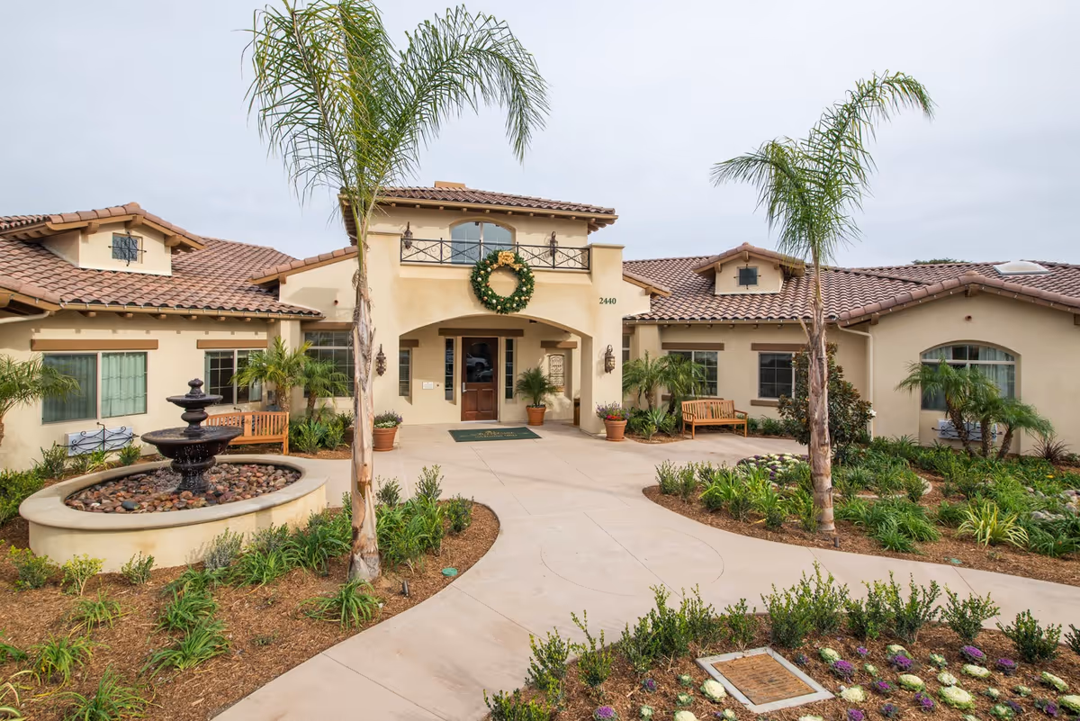 Exterior view of a single-story building with a tiled roof, featuring a central entrance adorned with a large green wreath. The entrance is flanked by potted plants and benches, with a circular fountain surrounded by landscaping and palm trees in the foreground.