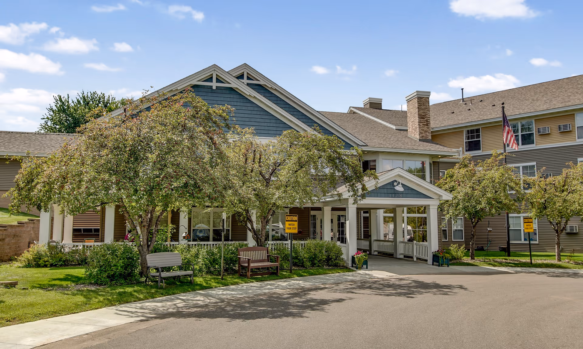 Exterior view of Oak Ridge Assisted Living facility showing a large building with multiple windows, a covered entrance with white pillars, trees, benches, and a clear blue sky.
