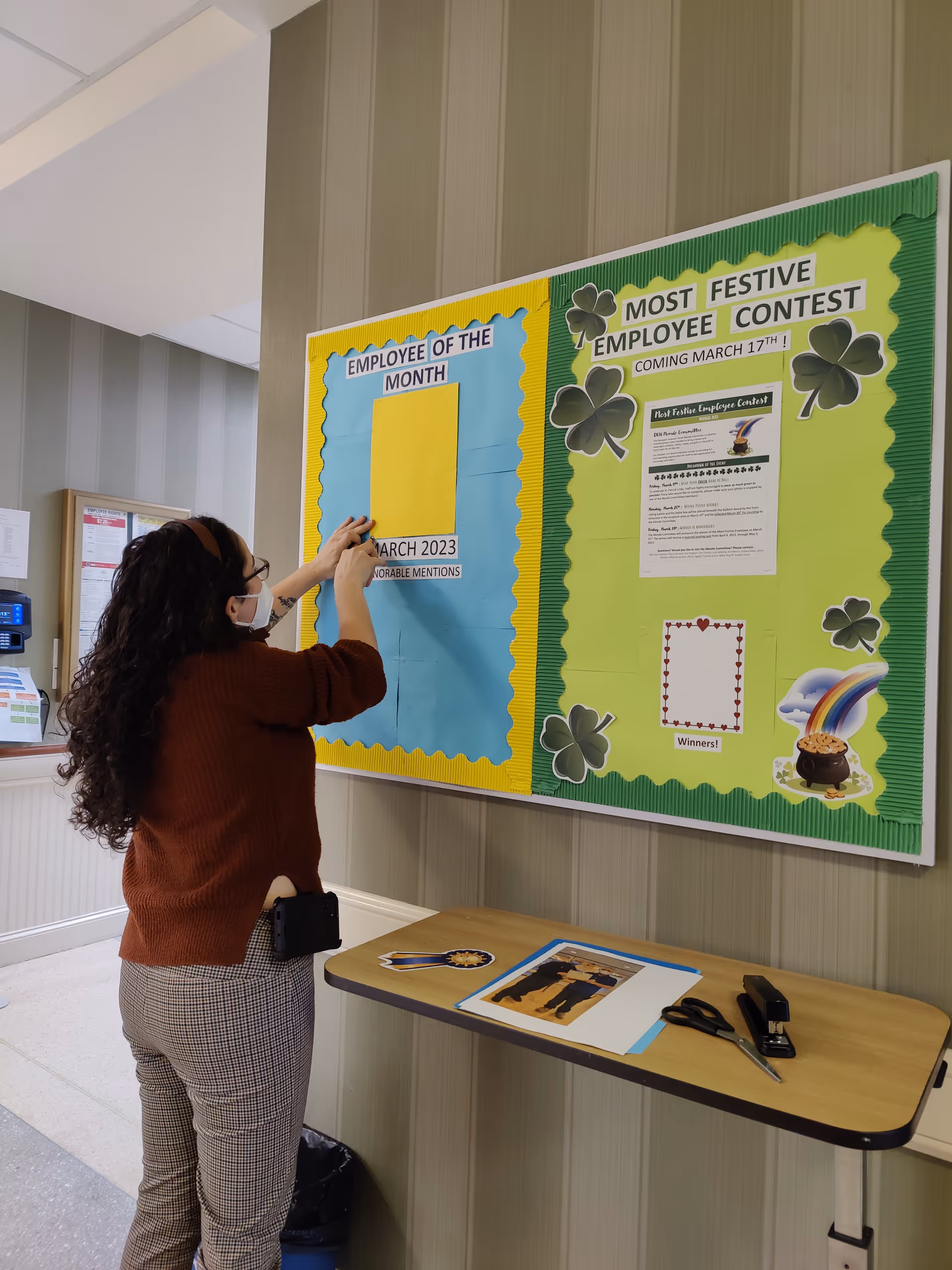 A woman wearing a mask and glasses is putting up a photo or paper on a bulletin board inside a facility. The bulletin board has two sections: one titled 'Employee of the Month' with a yellow and blue background, and the other titled 'Most Festive Employee Contest' with a green background decorated with shamrocks and a rainbow with a pot of gold. A small table below the board holds scissors, a stapler, and some papers.