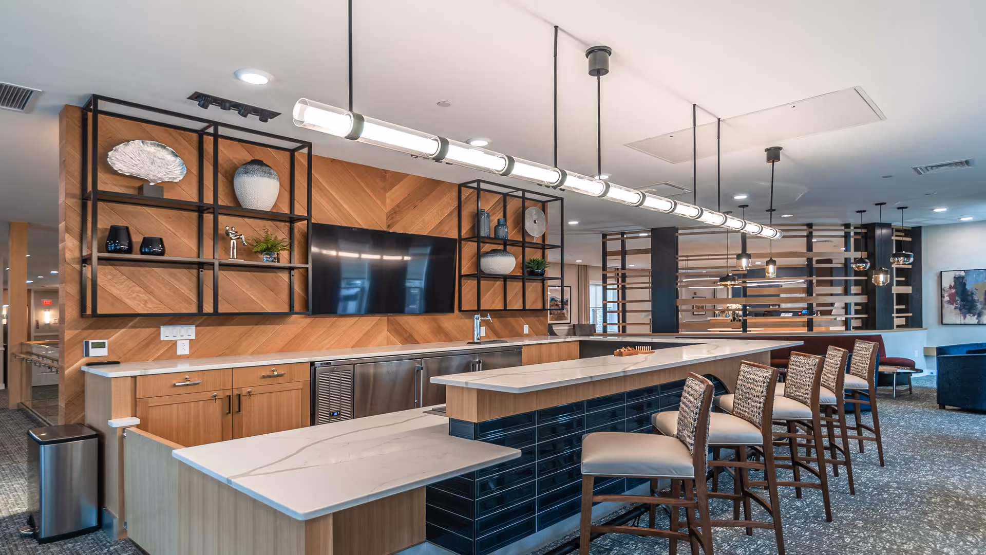 Modern interior space with a long marble countertop bar with five high chairs. Behind the bar is a wooden wall with decorative shelves holding vases and plants, and a mounted flat-screen TV. Pendant lights hang from the ceiling, and the area extends into a lounge with additional seating and artwork on the walls.