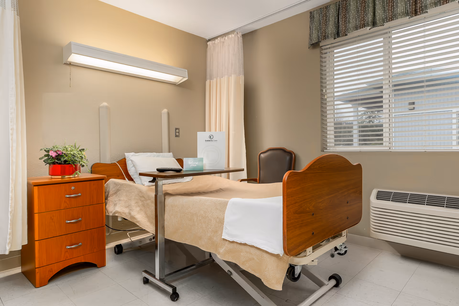 A hospital-style bed with beige bedding and a white pillow in a room with beige walls. Next to the bed is a wooden nightstand with three drawers and a potted plant on top. A small overbed table is positioned over the bed, holding a remote control and some pamphlets. There is a window with blinds and a valance, and a heating/cooling unit below the window. A single chair is placed against the wall near the window.