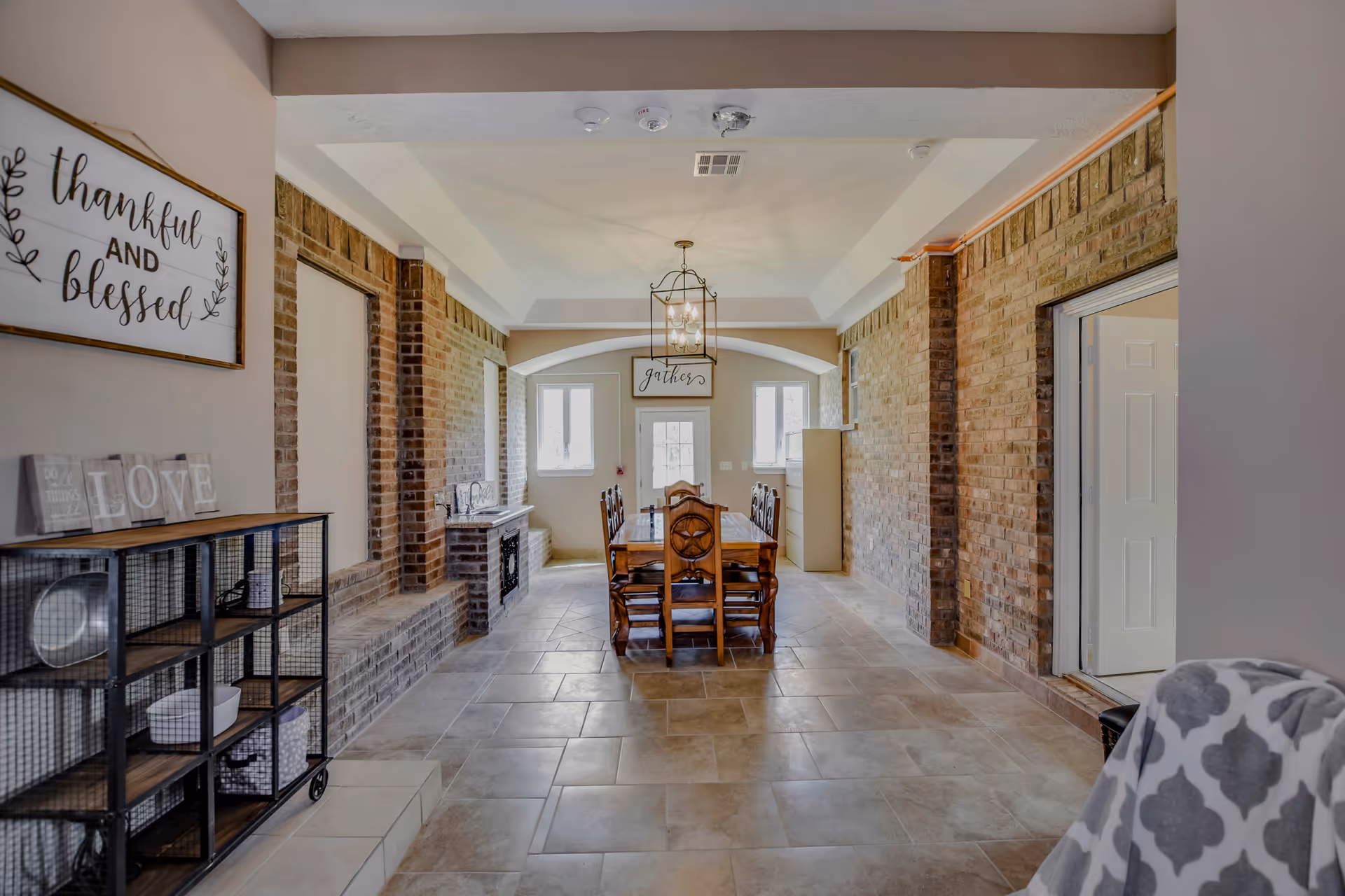 Long dining room with a wooden table and chairs centered under a chandelier, flanked by exposed brick walls and decorative signs.