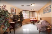 A cozy living room with a floral-patterned sofa, a small round table with two chairs near a window, a wooden TV stand with a television, and two large potted plants. The walls are light-colored with wood paneling on the lower half, and there are framed paintings hanging on the walls.